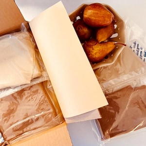May include: An open cardboard box containing packaged food items. Inside are sealed bags of ingredients, a basket of cooked pears, and a printed sheet with text. The box also includes a round sticker with text.