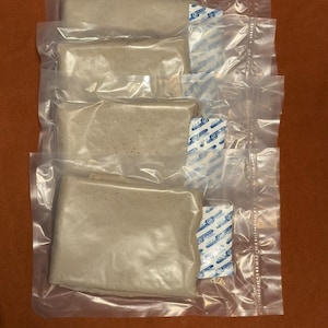 May include: Four individually packaged, rectangular food items in clear plastic. Each package contains a beige-colored food product and a small, blue and white packet. The packages are stacked on a brown surface.
