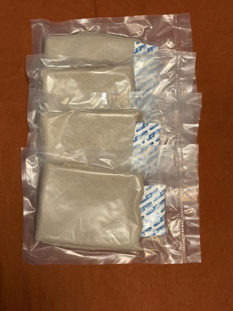 May include: Four individually packaged, rectangular food items in clear plastic. Each package contains a beige-colored food product and a small, blue and white packet. The packages are stacked on a brown surface.