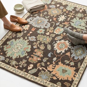 Peut inclure: Un tapis marron fonc&eacute; avec un motif floral dans des tons de bleu, vert et p&ecirc;che. Le tapis a une bordure beige et est pos&eacute; sur une surface blanche. Une couverture beige pli&eacute;e, de petits bols et des mocassins en cuir marron sont &agrave; c&ocirc;t&eacute; du tapis.