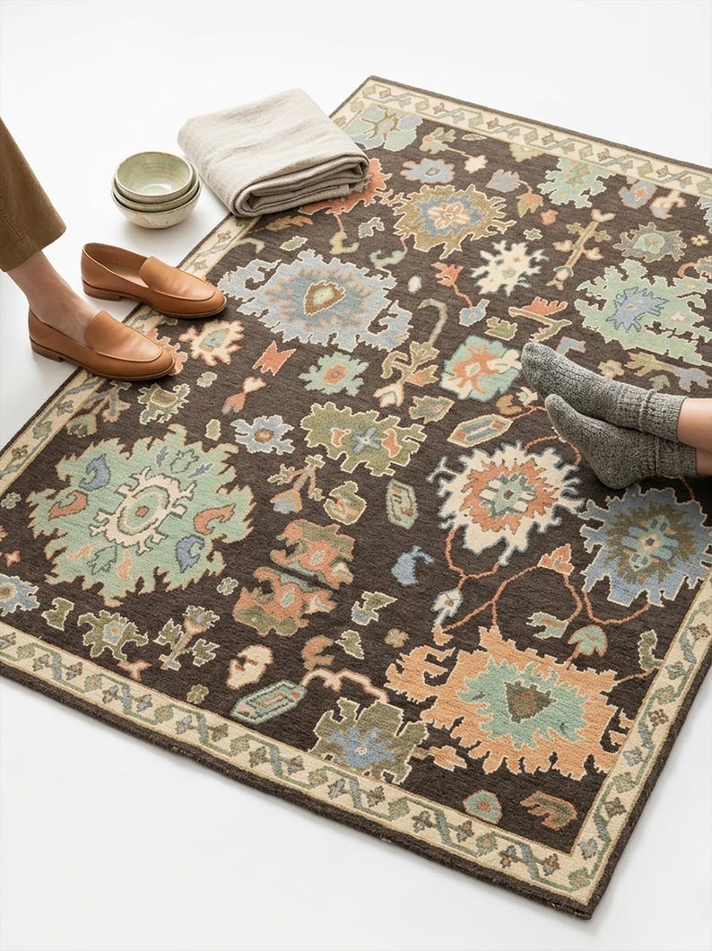Peut inclure: Un tapis marron fonc&eacute; avec un motif floral dans des tons de bleu, vert et p&ecirc;che. Le tapis a une bordure beige et est pos&eacute; sur une surface blanche. Une couverture beige pli&eacute;e, de petits bols et des mocassins en cuir marron sont &agrave; c&ocirc;t&eacute; du tapis.