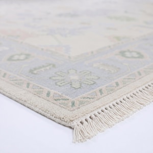 May include: A rectangular area rug featuring a cream-colored center with soft floral patterns in muted blue, green, and pink. The rug has a wide border with geometric and floral motifs, finished with a short, cream-colored fringe along the edge.