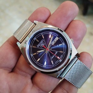 May include: A silver-toned Seiko automatic wristwatch with a mesh band. The watch face is a deep blue with white and red accents, including the day and date. The watch is marked "Japan Made".