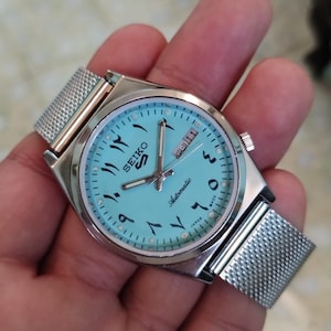 Puede incluir: Un reloj automático Seiko 5 plateado con esfera azul claro y números arábigos. El reloj tiene una correa de malla y una pantalla de fecha. La esfera del reloj también tiene la palabra "Automatic" y "Japan".