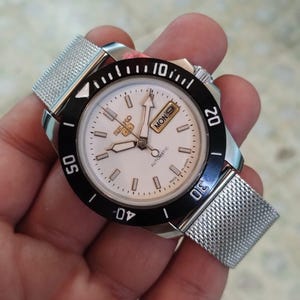 May include: A silver-toned Seiko 5 automatic watch with a white face, black bezel, and mesh band. The watch face displays the day and date. The bezel has white markings. The watch is held in a hand.