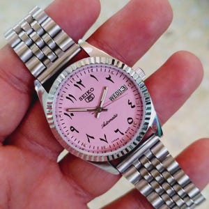 May include: A silver Seiko 5 automatic watch with a pink face and Arabic numerals. The watch has a day and date display and a stainless steel strap. The watch is held in a hand.