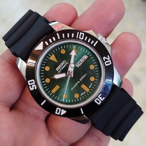 Può includere: Un orologio subacqueo Seiko Automatic in acciaio inossidabile con lunetta nera e quadrante verde. L'orologio presenta un cinturino in gomma nera e mostra il giorno e la data. Il quadrante ha indici e lancette luminescenti.