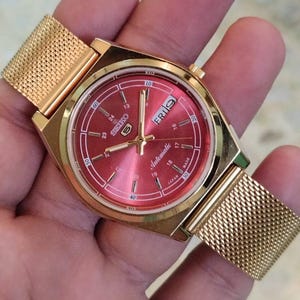 Peut inclure: Une montre automatique Seiko 5 dorée avec un cadran rouge et un bracelet en maille. Le cadran affiche le jour et la date, avec le mot "Automatic" visible. La montre est tenue dans une main.