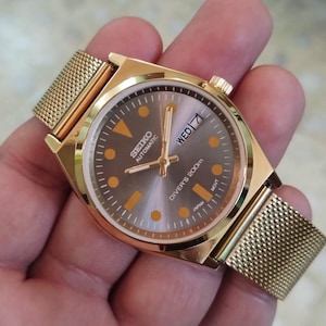 Op de afbeelding: Goudkleurig Seiko automatisch horloge met een mesh band. De wijzerplaat van het horloge is grijs met oranje markeringen en wijzers. De dag en datum worden op 3 uur weergegeven. Het horloge is gemarkeerd met "Diver's 200m" en "Japan".