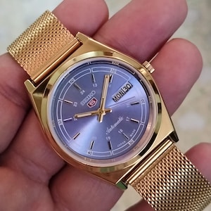 Puede incluir: Un reloj automático Seiko 5 dorado con una correa de malla. La esfera del reloj es de color azul claro con agujas y marcadores dorados. El día y la fecha se muestran a las 3 en punto. El reloj se sostiene en una mano.
