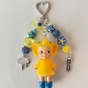 May include: A quirky keychain with a yellow doll wearing blue boots and a matching yellow outfit. The keychain has a silver heart-shaped clasp, a beaded strand with blue and yellow details, and charms including a cloud and a vintage phone.