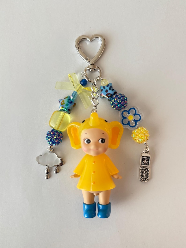 May include: A quirky keychain with a yellow doll wearing blue boots and a matching yellow outfit. The keychain has a silver heart-shaped clasp, a beaded strand with blue and yellow details, and charms including a cloud and a vintage phone.