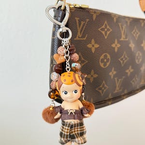K&ouml;nnte beinhalten: Ein dekorativer Schl&uuml;sselanh&auml;nger mit einer kleinen Puppe in einem braunen und orangefarbenen Outfit, braunen Haaren und einem gelben Hut. Der Schl&uuml;sselanh&auml;nger enth&auml;lt braune und goldene Perlen und braune Fellakzente. Er ist an einer braunen Handtasche mit einer goldenen Kette befestigt.