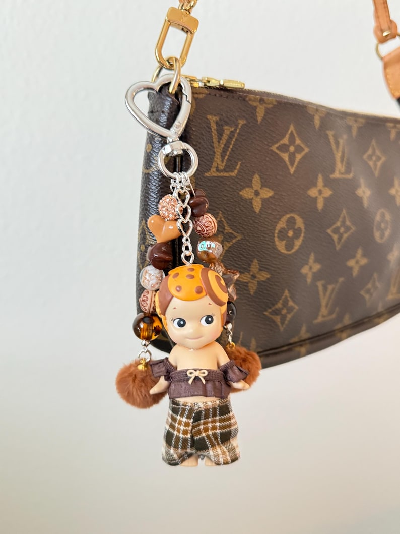 K&ouml;nnte beinhalten: Ein dekorativer Schl&uuml;sselanh&auml;nger mit einer kleinen Puppe in einem braunen und orangefarbenen Outfit, braunen Haaren und einem gelben Hut. Der Schl&uuml;sselanh&auml;nger enth&auml;lt braune und goldene Perlen und braune Fellakzente. Er ist an einer braunen Handtasche mit einer goldenen Kette befestigt.