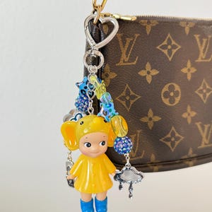 May include: A quirky keychain charm with a yellow figure in a raincoat and blue wellington boots, decorated with beaded details and a silver charm. The charm is attached to a gold-toned clasp, with a brown and tan handbag in the background.