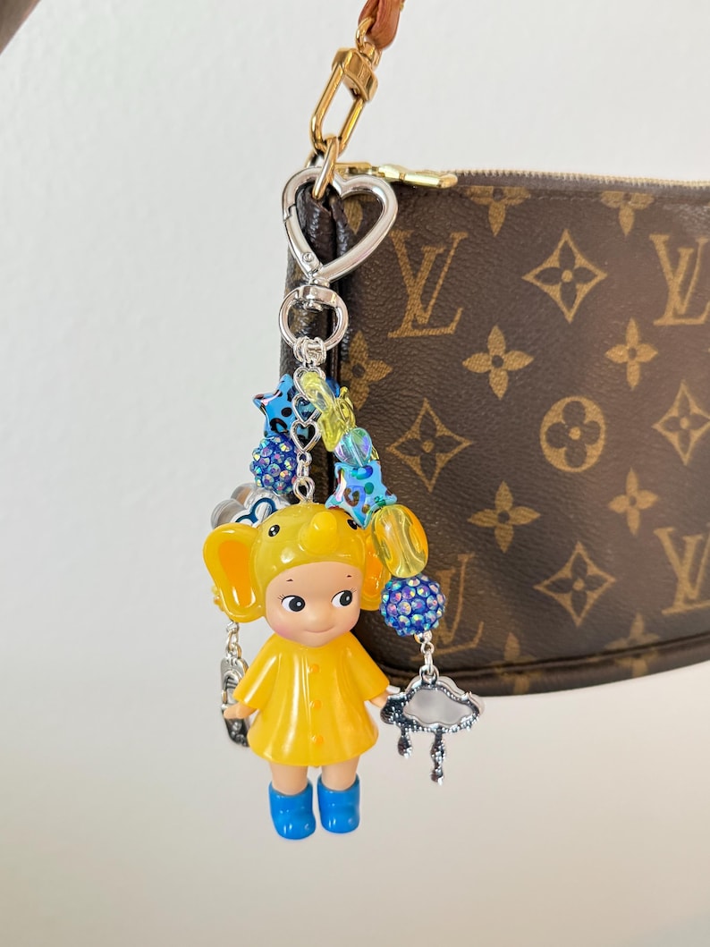 May include: A quirky keychain charm with a yellow figure in a raincoat and blue wellington boots, decorated with beaded details and a silver charm. The charm is attached to a gold-toned clasp, with a brown and tan handbag in the background.