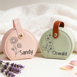 Könnte beinhalten: Zwei kleine, runde, personalisierte Schmuckkästchen. Eines ist hellrosa mit einem Gänseblümchen-Design und dem Namen "Sandy". Das andere ist hellgrün mit einem Blumendesign und dem Namen "Oswald". Beide haben einen Griff und goldfarbene Beschläge.