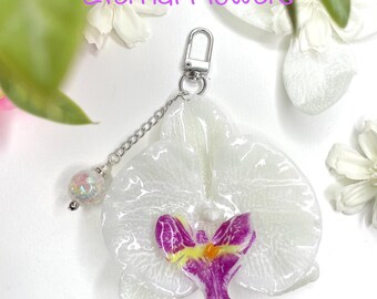 May include: A white orchid flower charm with a silver chain and clasp. The flower has a purple and yellow centre. The charm is attached to a silver chain with a decorative iridescent bead. The text "Eternal Flowers" is visible.