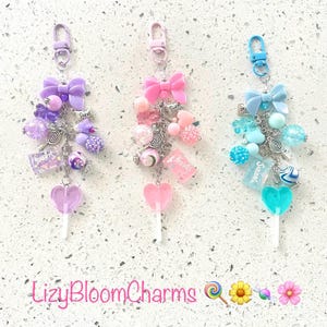 May include: Three charm keyrings with bows, beads, and lollipop pendants. The keyrings feature purple, pink, and blue colour schemes, with various bead shapes and sizes. Each keyring has a lobster clasp for easy attachment.