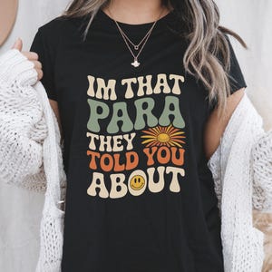 K&ouml;nnte beinhalten: Schwarzes T-Shirt mit dem Text "IM THAT PARA THEY TOLD YOU ABOUT" in Retro-Schrift. Das Design beinhaltet eine G&auml;nsebl&uuml;mchen und ein Smiley-Gesicht. Es ist ein Freizeitkleidungsst&uuml;ck.