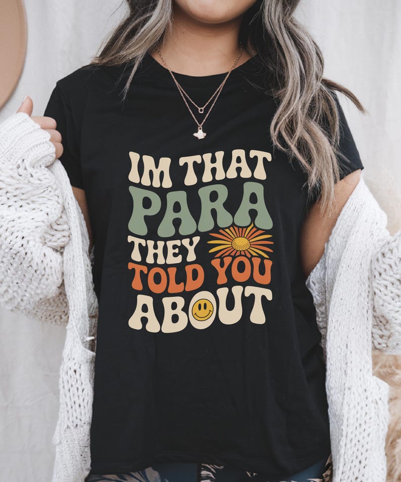 K&ouml;nnte beinhalten: Schwarzes T-Shirt mit dem Text "IM THAT PARA THEY TOLD YOU ABOUT" in Retro-Schrift. Das Design beinhaltet eine G&auml;nsebl&uuml;mchen und ein Smiley-Gesicht. Es ist ein Freizeitkleidungsst&uuml;ck.