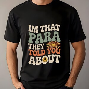 K&ouml;nnte beinhalten: Schwarzes T-Shirt mit dem Text "IM THAT PARA THEY TOLD YOU ABOUT" in einem Retro-Schriftstil. Das Design beinhaltet eine G&auml;nsebl&uuml;mchen und ein Smiley-Gesicht. Das T-Shirt hat einen Rundhalsausschnitt.