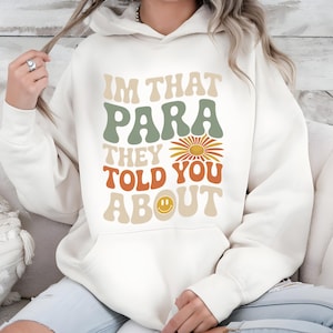 K&ouml;nnte beinhalten: Cremefarbener Kapuzenpullover mit dem Text "IM THAT PARA THEY TOLD YOU ABOUT" in Retro-Schrift. Das Design beinhaltet eine G&auml;nsebl&uuml;mchen und ein Smiley-Gesicht. Der Hoodie hat eine Vordertasche und eine Kapuze mit Kordelzug.