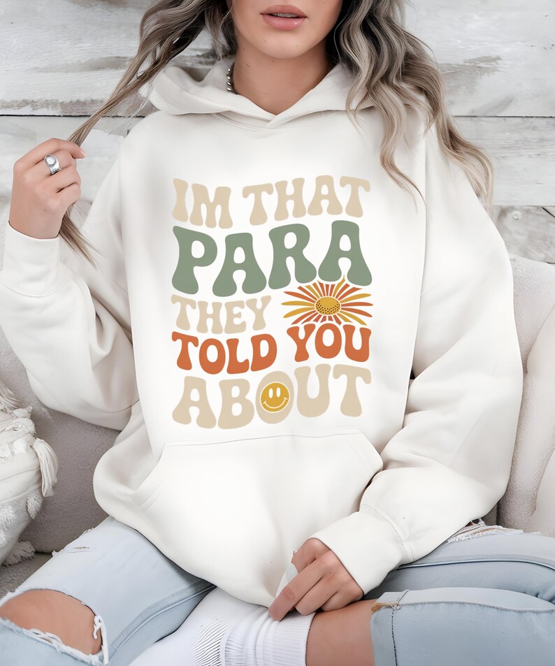 K&ouml;nnte beinhalten: Cremefarbener Kapuzenpullover mit dem Text "IM THAT PARA THEY TOLD YOU ABOUT" in Retro-Schrift. Das Design beinhaltet eine G&auml;nsebl&uuml;mchen und ein Smiley-Gesicht. Der Hoodie hat eine Vordertasche und eine Kapuze mit Kordelzug.
