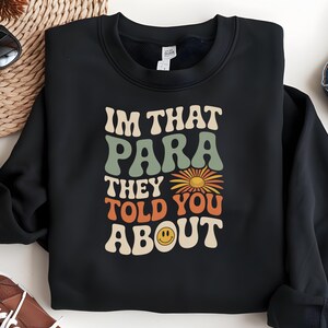 K&ouml;nnte beinhalten: Schwarzes Sweatshirt mit dem Text "I'M THAT PARA THEY TOLD YOU ABOUT" in Retro-Schrift. Das Design beinhaltet eine Sonnenblume und ein Smiley-Gesicht. Das Sweatshirt ist auf einer hellen Oberfl&auml;che gefaltet.