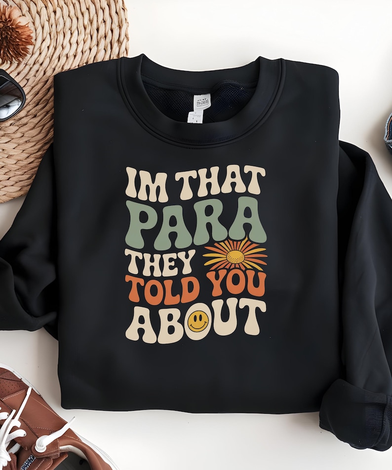 K&ouml;nnte beinhalten: Schwarzes Sweatshirt mit dem Text "I'M THAT PARA THEY TOLD YOU ABOUT" in Retro-Schrift. Das Design beinhaltet eine Sonnenblume und ein Smiley-Gesicht. Das Sweatshirt ist auf einer hellen Oberfl&auml;che gefaltet.