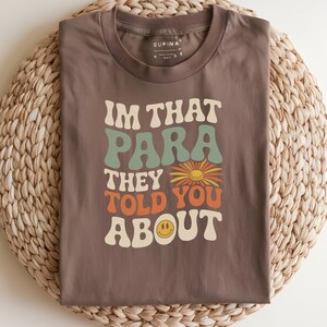 K&ouml;nnte beinhalten: Braunes T-Shirt mit dem Text "IM THAT PARA THEY TOLD YOU ABOUT" in Retro-Schrift. Das Design beinhaltet eine Blume und ein l&auml;chelndes Ei. Das T-Shirt liegt gefaltet auf einer gewebten Matte.