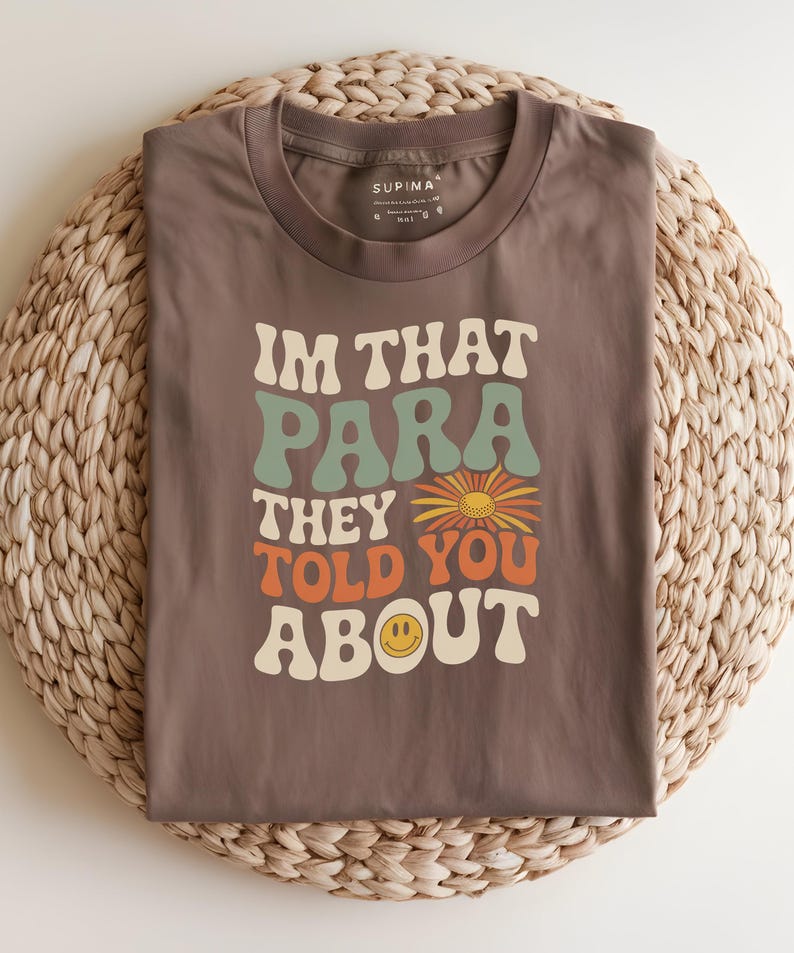 K&ouml;nnte beinhalten: Braunes T-Shirt mit dem Text "IM THAT PARA THEY TOLD YOU ABOUT" in Retro-Schrift. Das Design beinhaltet eine Blume und ein l&auml;chelndes Ei. Das T-Shirt liegt gefaltet auf einer gewebten Matte.