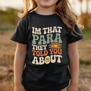 K&ouml;nnte beinhalten: Schwarzes T-Shirt mit dem Text "IM THAT PARA THEY TOLD YOU ABOUT" in Retro-Schrift. Das Design beinhaltet eine Blume und ein Smiley. Das T-Shirt wird von einem Kind getragen.