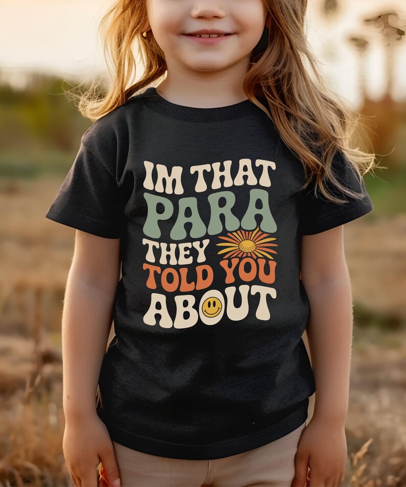 K&ouml;nnte beinhalten: Schwarzes T-Shirt mit dem Text "IM THAT PARA THEY TOLD YOU ABOUT" in Retro-Schrift. Das Design beinhaltet eine Blume und ein Smiley. Das T-Shirt wird von einem Kind getragen.