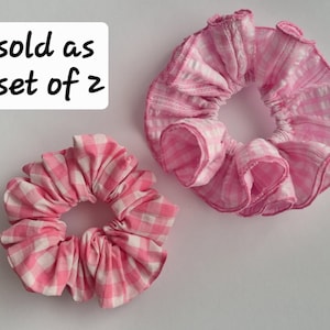 Könnte beinhalten: Zwei rosa-weiß karierte Haargummis. Die Haargummis sind aus einem gerüschten Stoff gefertigt und werden auf weißem Hintergrund gezeigt. Der Text "sold as set of 2" befindet sich in einem weißen Feld.