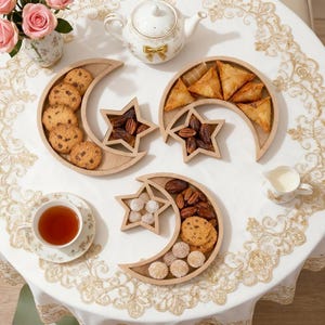 May include: A table set with crescent and star-shaped wooden serving platters, laden with biscuits, pastries, and dates. A teapot, teacup, and creamer are also present, alongside a vase of pink roses.