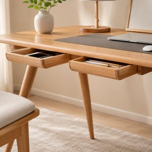 May include: A light-toned wooden desk featuring two shallow, open drawers positioned side-by-side under the tabletop. The desk surface holds a dark grey desk mat, a laptop, a white computer mouse, a wooden lamp base, and a white ceramic vase.
