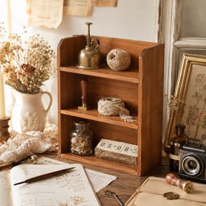 May include: A three-tiered wooden shelf unit featuring a brass oil can and a ball of twine on the top shelf, a wax seal stamp and lace ribbon on the middle shelf, and a glass jar with contents and a wooden block calendar on the bottom shelf.