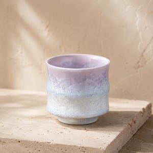May include: A ceramic cup featuring a gradient colour transition from a soft lavender at the rim to a pale blue and white at the base. The surface has a distinct crystalline, crackled glaze texture that creates a multifaceted, reflective appearance.