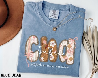 May include: A blue jean colored t-shirt featuring the name Isabel in large, patterned letters with floral and checkered designs. The text certified nursing assistant is printed below in white, accented by small pink bows on the letters.