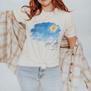 Puede incluir: Una camiseta de manga corta color crema con un gráfico estilo acuarela de un cielo azul con un sol amarillo. El texto dice LESS CLOUD. More Sky DEFEAT AI. La camiseta se lleva con una camisa de cuadros beige y blanca y unos vaqueros azul claro.
