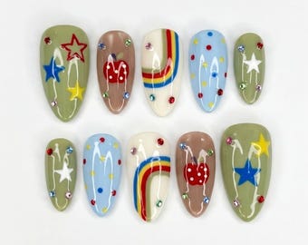 May include: A set of ten artificial nails in various designs. The nails feature a mix of colors, including olive green, brown, white, and blue. Designs include stars, apples, rainbows, and polka dots, embellished with small, colorful rhinestones.