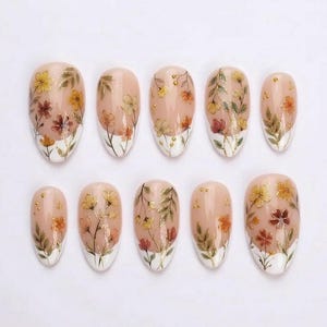 Può includere: Un set di dieci unghie finte ovali con una base color nude. Ogni unghia presenta un delicato disegno floreale dipinto a mano nei toni del giallo, arancione e marrone, accentuato da sottili steli verdi e piccoli punti metallici dorati.