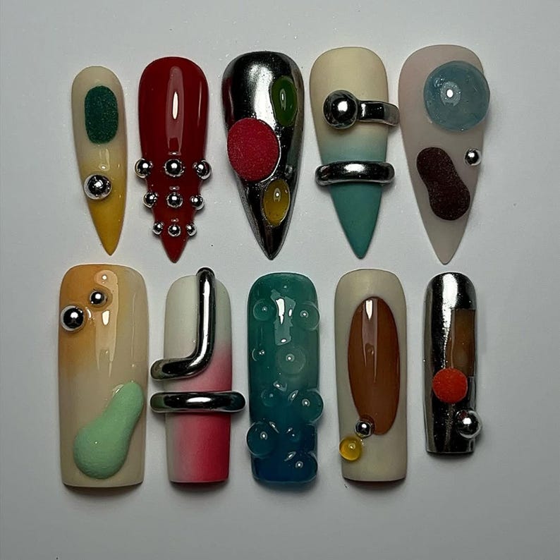 May include: A collection of ten uniquely designed artificial nails. Each nail features a different design, including metallic accents, colorful embellishments, and abstract shapes. The nails showcase a variety of colors, including red, green, silver, and beige.
