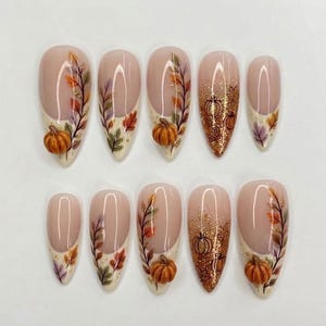 Può includere: Un set di dieci unghie finte a mandorla con base color carne e decorazioni a tema autunnale. Le unghie presentano foglie dipinte a mano in arancione, giallo e viola, accenti di glitter dorato e piccole zucche tridimensionali in rilievo.