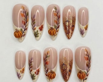 May include: A set of ten almond-shaped press-on nails featuring a nude base with autumn-themed designs. The nails include hand-painted orange, yellow, and purple leaves, gold glitter accents, and small three-dimensional orange pumpkin charms.