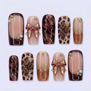 Op de afbeelding: Een set van tien kunstnagels met diverse ontwerpen, waaronder bruine French tips met parels, metallic gouden strikken, bruin-zwarte marmeren wervelingen, luipaardprint met koffieboonaccenten en glinsterende oranje pompoenen met gouden omlijningen.