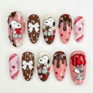 May include: A set of ten almond-shaped nail designs featuring Snoopy characters and Valentine's Day themes. Designs include Snoopy holding hearts, chocolate drips, bows, and candy cane stripes, with pink, white, and brown colors.