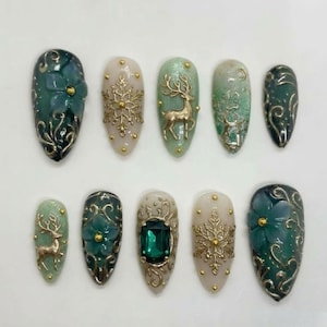 May include: A set of ten almond-shaped press-on nails featuring a festive theme. The designs include dark green and cream backgrounds adorned with gold metallic filigree, raised snowflake motifs, deer silhouettes, and green floral accents.