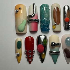 May include: A collection of uniquely designed artificial nails. Each nail features a different design, including metallic accents, textured elements, and various color gradients. The nails are in different shapes and sizes, showcasing a variety of artistic styles.